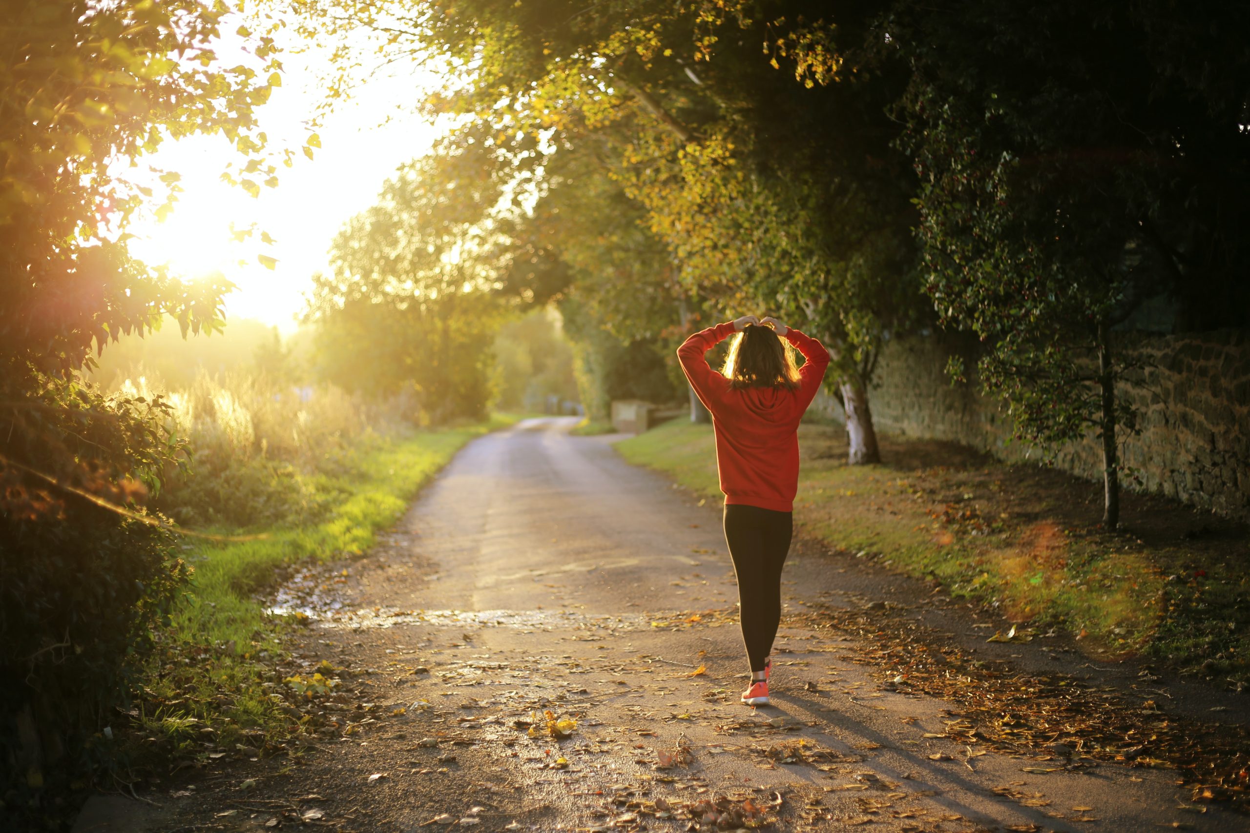 Woman in a red shirt and black pants walking on a gravel trail in the woods at sunset