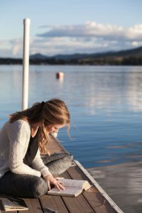 Woman reading a book on a dock near a lake.